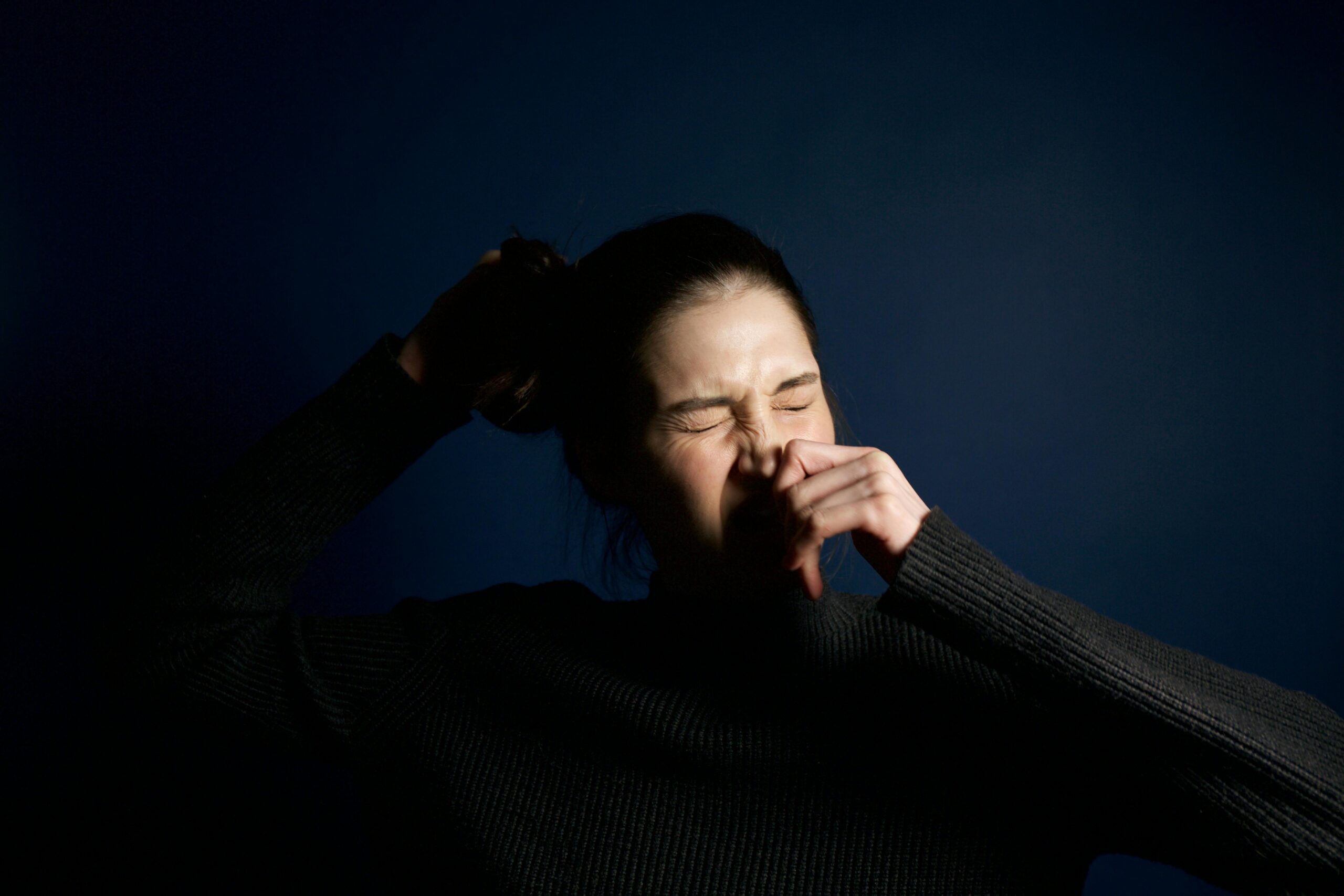 A woman in a dark sweater sneezes against a solid blue background, capturing a moment of candid expression.